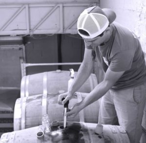 Une personne portant une casquette de baseball à l'envers utilise un outil pour extraire le FÛT DE BIÈRE BLONDE 5° 30L (Bière artisanale bouledogue) d'un tonneau en bois dans un environnement industriel, avec plusieurs tonneaux et petits récipients en verre visibles.