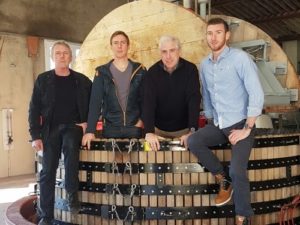 Quatre hommes posent près d'un grand pressoir en bois dans une cave industrielle. Habillés de façon décontractée, ils se tiennent debout ou assis avec un MAGNUM DE CHAMPAGNE ANDRÉ DILIGENT (Brut Blanc de blanc 150cl), en regardant l'appareil photo.