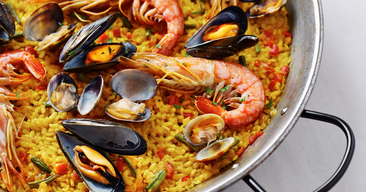Gros plan sur une paella traditionnelle aux fruits de mer, composée de riz jaune, de crevettes, de moules, de palourdes et de légumes, servie dans une grande poêle ronde à poignées métalliques.
